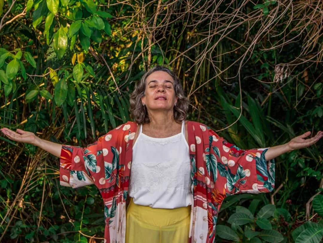Aurì Filha em jardim natural, postura de acolhimento com braços abertos, expressão serena